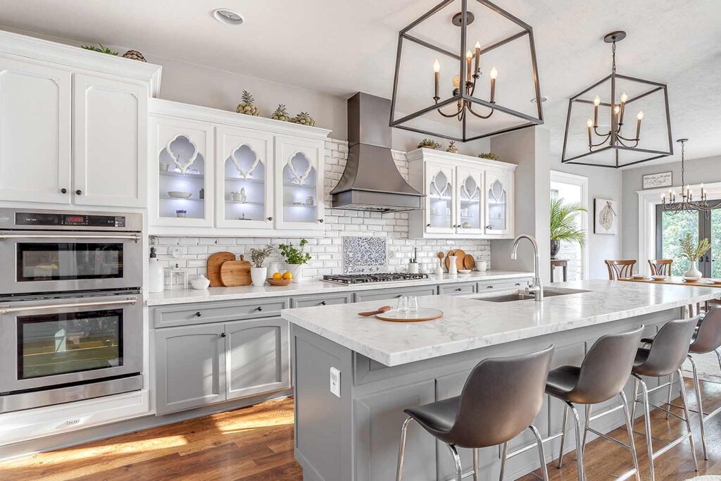 Classic two-toned kitchen in a Scottsdale neighborhood featuring soft muted gray recessed panel lower cabinets and white upper cabinets with decorative glass fronts. The kitchen has freshly painted walls in light gray, shiny white trim, and medium-tone herringbone wood flooring. Counters are styled with wooden cutting boards, ceramic jars, fresh fruit, and hanging kitchen utensils. Harsh late morning sunlight streams through a large black-framed window casting dramatic shadows and bright highlights, creating a moody and inviting atmosphere, gray and white kitchen, average cabinet painting cost in Phoenix Arizona, Average Cost to paint cabinets Phoenix Arcadia AZ, cost to paint cabinets, cabinet painting cost, cabinet painting phoenix, painting cabinets vs replacing them, cost to replace cabinets, two tone cabinets