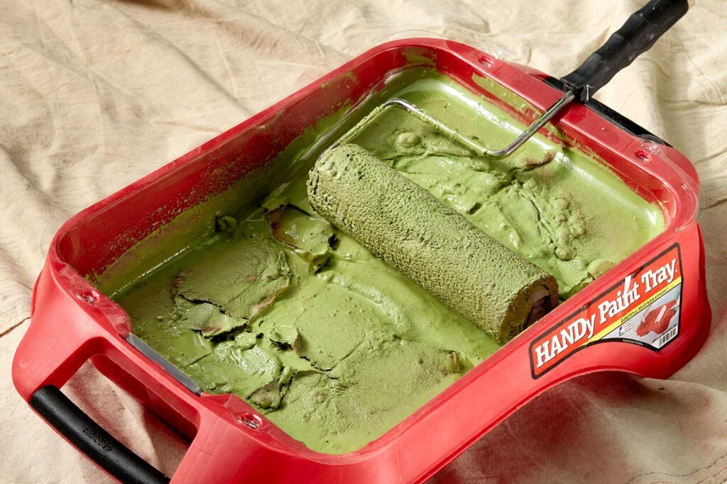 Paint Tray and Paint Roller with Skinning Due to Hot Weather, Close-up of a red HANDy Paint Tray filled with thick green paint and a paint roller resting inside, set on a beige drop cloth, showing texture and consistency of the paint.