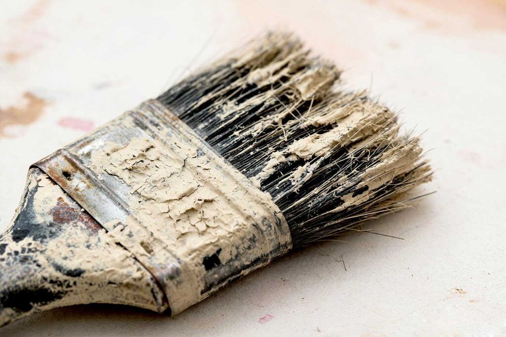 Paintbrush with Dried Paint on Heel and Bristles, Close-up of an old, worn paintbrush with stiff, cracked beige paint on the bristles and metal ferrule, resting on a white surface, illustrating heavy use and dried paint buildup.