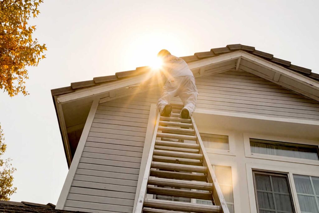 Painter on a Ladder in Blistering Heat, A man wearing white painter’s overalls climbing a tall aluminum ladder to reach the roofline of a house, with the sun shining brightly behind, casting a warm glow.