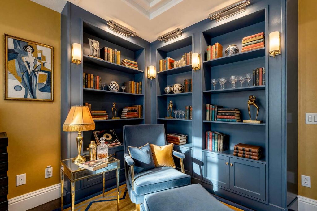 Art Deco Reading Corner with Slate Blue Cabinets and Golden Ochre Walls-3, elegant art deco reading corner slate blue cabinets golden ochre accent wall soft off-white ceiling crisp white trim. A refined reading nook in a Scottsdale neighborhood featuring slate blue built-in bookshelves against a golden ochre accent wall, perfect for painting built-in bookshelves with bold bookshelf paint color ideas. Velvet armchair, mirrored side table, and geometric pottery create a glamorous 1920s vibe. Morning sunlight highlights the rich colors and sophisticated details. paint colors for built-in shelving, built-in cabinetry paint ideas, bookshelf color palette ideas, painted bookcase design inspiration, colors for built-in bookshelves, bookshelf and cabinet paint pairings