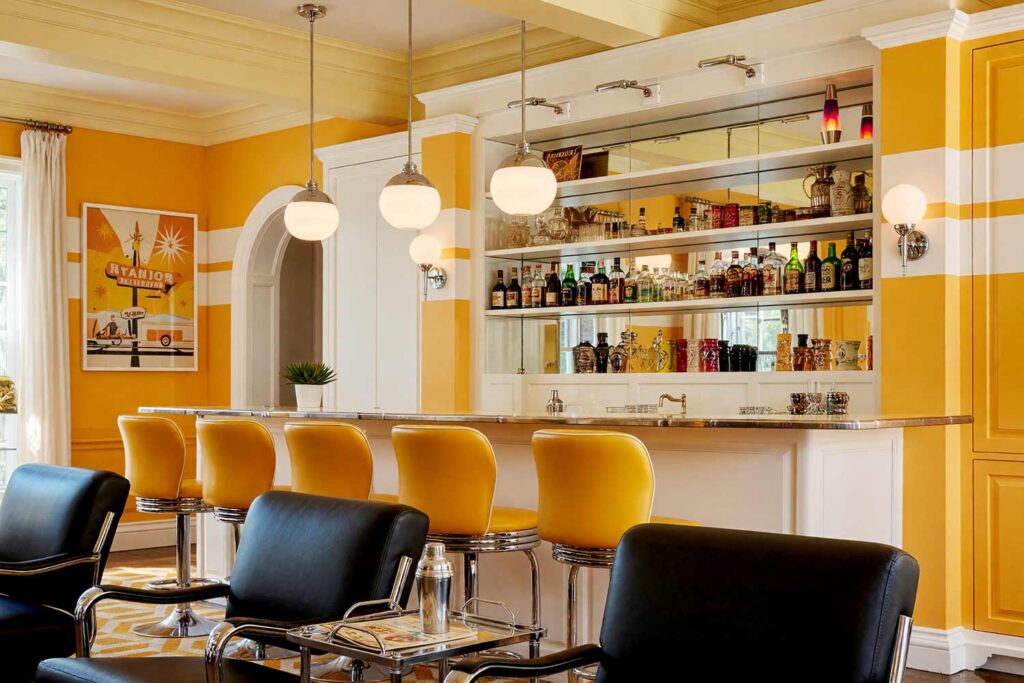 Fun Retro Atomic Home Bar with Marigold and White Striped Walls-6, fun retro atomic home bar warm marigold soft white striped walls pale yellow ceiling bright white trim. A nostalgic home bar in a Scottsdale neighborhood featuring bold marigold and white striped walls, soft white cabinetry, and chrome accents. Décor includes retro diner pop art, lava lamps, tiki mugs, and chrome pendant lighting. Dramatic natural and artificial light highlights the playful mid-century modern palette and textures of this lively space. bar cabinet paint colors, home bar color palette, wine nook paint colors, built-in bar paint scheme, bar area color combinations