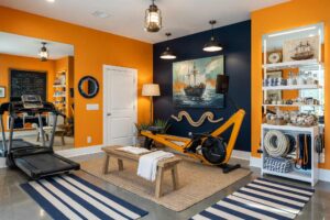 Nautical Rowing Sanctuary with Marigold Walls Navy Accent and White Trim-4-FEATURE, nautical home gym paint colors marigold orange walls navy ink accent wall bright white trim polished concrete flooring rowing machine scottsdale. A premium rowing sanctuary in a Scottsdale neighborhood featuring a matte black Concept2 RowErg positioned against a dramatic navy accent wall, surrounded by vibrant marigold energy. Curated maritime décor includes a coiled rope wall sculpture, a painted porthole mirror, floating shelves displaying nautical charts and brass compasses, and a briny ocean-air diffuser creating an immersive coastal atmosphere. Polished concrete flooring and bright white trim frame the space with precision, while dramatic natural light and brass ship's lanterns create dynamic shadows and highlights. Every element reinforces the sensation of cutting through open water—a luxurious, focused training environment that combines maritime swagger with serious athletic purpose. home gym paint colors, gym paint colors, workout room color, home gym color palette, home gym color scheme, workout space paint colors, exercise room paint colors