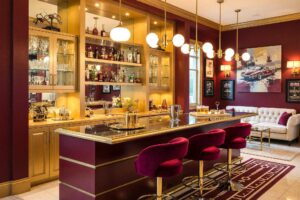 Racing Home Bar with Burgundy Walls Gold Millwork and Cream Ceiling-1-FEATURE, luxurious home bar burgundy walls gold millwork cream ceiling deep burgundy trim 1950s design. A glamorous home bar in a Scottsdale neighborhood featuring a curved gold bar with burgundy velvet stools, gold-accented lighting, and racing-inspired artwork. Built-in shelves display liquor and memorabilia, while dramatic lighting enhances the rich, vintage palette of this sophisticated space. home bar paint colors, house paint colors, paint color inspiration, bar nook colors, wet bar paint colors, cocktail nook color palette, entertainment nook paint scheme
