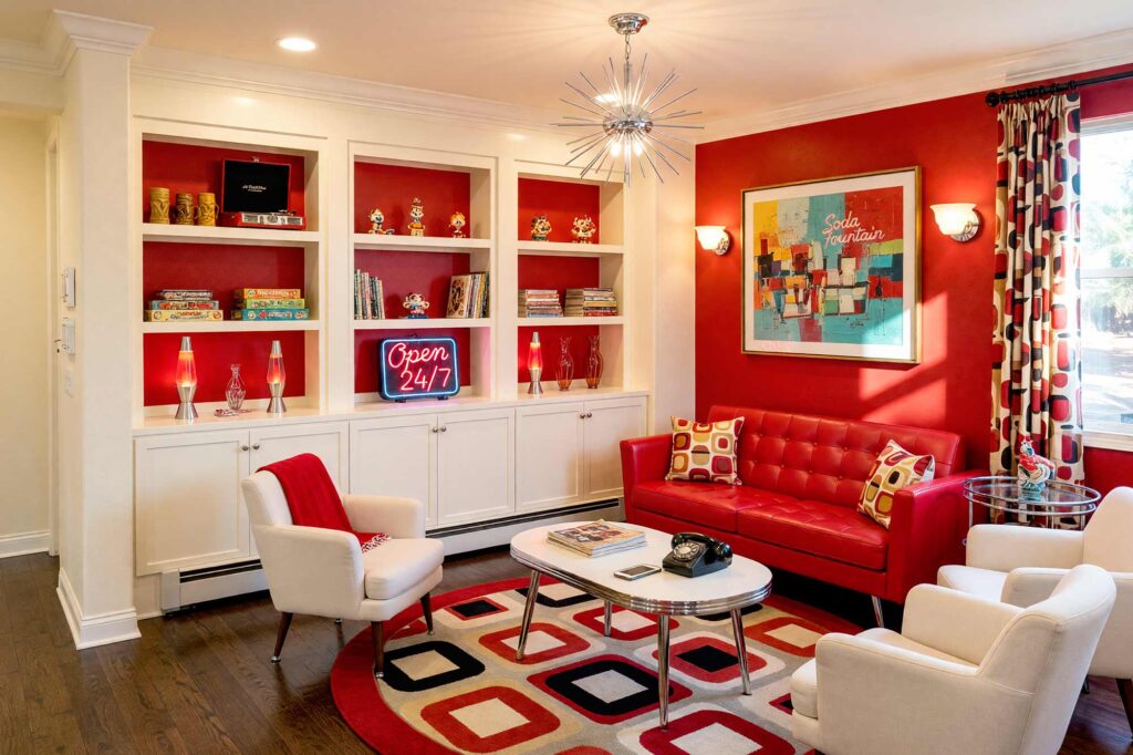 Retro Diner-Inspired Living Room with Cherry Red Accent Wall and Soft Cream Shelving-4-FEATURE, retro living room with cherry red accent wall soft cream shelving chrome trim and dark oak flooring. A vibrant 1950s-inspired living room in a Scottsdale neighborhood featuring a tufted cherry red vinyl sofa, kidney-shaped coffee table, and retro bar cart. Built-in shelving showcases vintage board games, tiki mugs, and a chrome-edged record player, while a starburst chandelier and bold geometric rug complete the nostalgic diner-style aesthetic. Bright morning sunlight casts dramatic shadows, enhancing the playful, glossy look of this charming mid-century space. Bookshelf paint color ideas, Painting built-in bookshelves, Color ideas for painting bookshelves, Built-in bookcase design ideas, Ideas for painting built-in shelves, Built-in bookshelf color inspiration, Bookcase paint colors