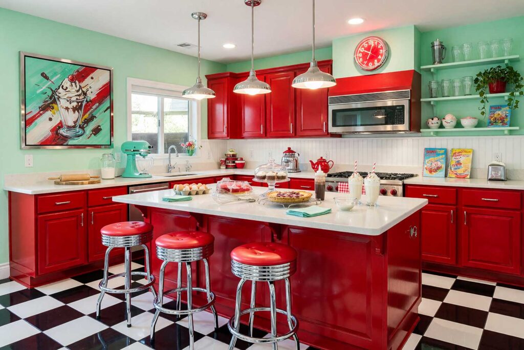 1950s Diner Kitchen with Mint Green Walls and Cherry Red Cabinets-3, kitsch themed room paint 1950s diner kitchen mint green walls cherry red cabinets crisp white ceiling checkerboard tile flooring. Located in an upscale Scottsdale neighborhood, this nostalgic culinary space features lustrous semi-gloss cherry red cabinets, chrome bar stools, crisp white trim, and mint green walls. Countertops are filled with authentic baking prep, from a cooling cherry pie to a vintage stand mixer, while strong natural daylight and a glowing neon clock bring bold retro drama to the dynamic diner aesthetic.