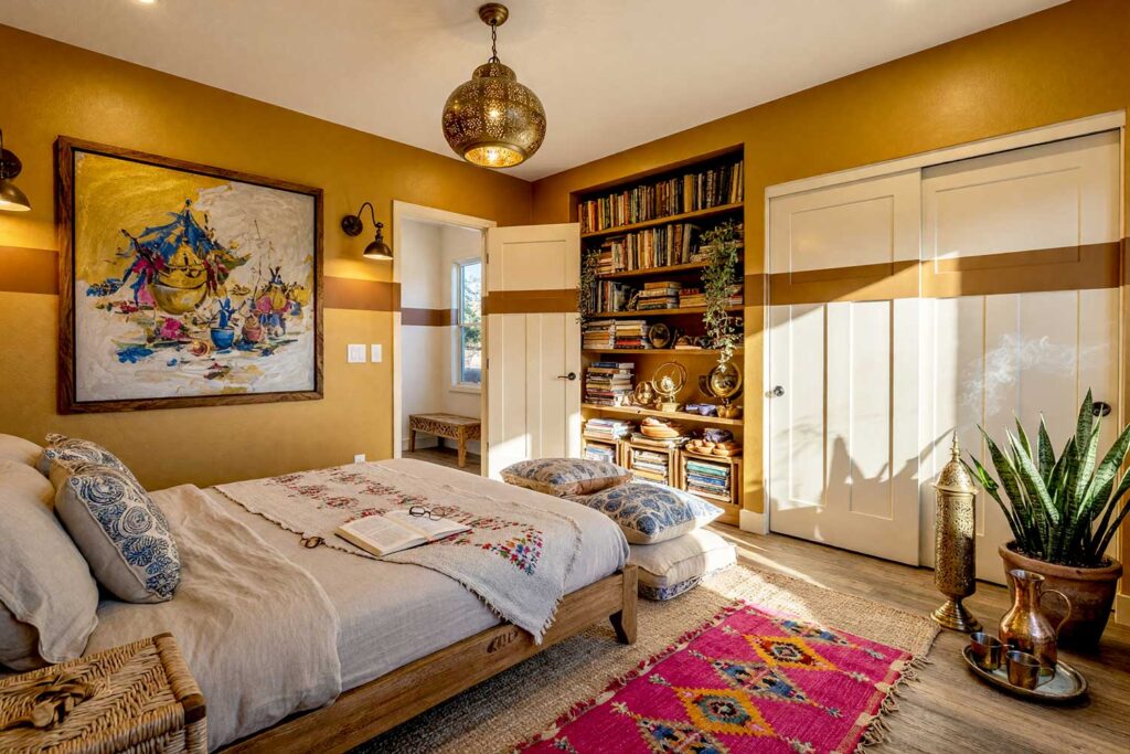 Bohemian Bedroom with Honey Gold Walls and Ochre Band-1, global bohemian bedroom honey gold walls and ochre painted wall graphics soft white ceiling distressed pine flooring. A captivating Scottsdale neighborhood retreat showcasing honey gold walls accented by a continuous horizontal ribbon band graphic in ochre. The room features a carved mango wood platform bed with rumpled linen bedding, rich multi-layered tribal rugs, a dense built-in bookcase filled with vintage travel books, and an evocative impressionist artwork. Strong directional afternoon light creates high-contrast shadows across the warm pine wood, delivering a tranquil and artistic bohemian sanctuary. painted wall graphics, wall murals, painted murals