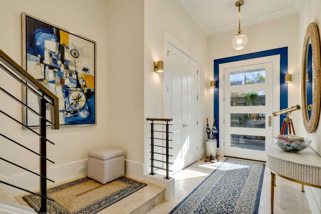 Entryway with Buttercream Walls and Cobalt Blue Door Halo-3, coastal entryway buttercream walls cobalt blue painted wall graphics cloud white ceiling travertine flooring. An inviting Scottsdale neighborhood foyer featuring bright buttercream walls, a cloud white ceiling, and a vibrant cobalt blue doorway halo graphic. Styled with a marble-topped console table, rope-framed mirror, authentic sea glass display, and dense built-in library shelving over polished travertine tiles. Strong morning light casts striking shadows across the space, delivering an elevated coastal resort look. painted wall graphics, wall murals, painted murals