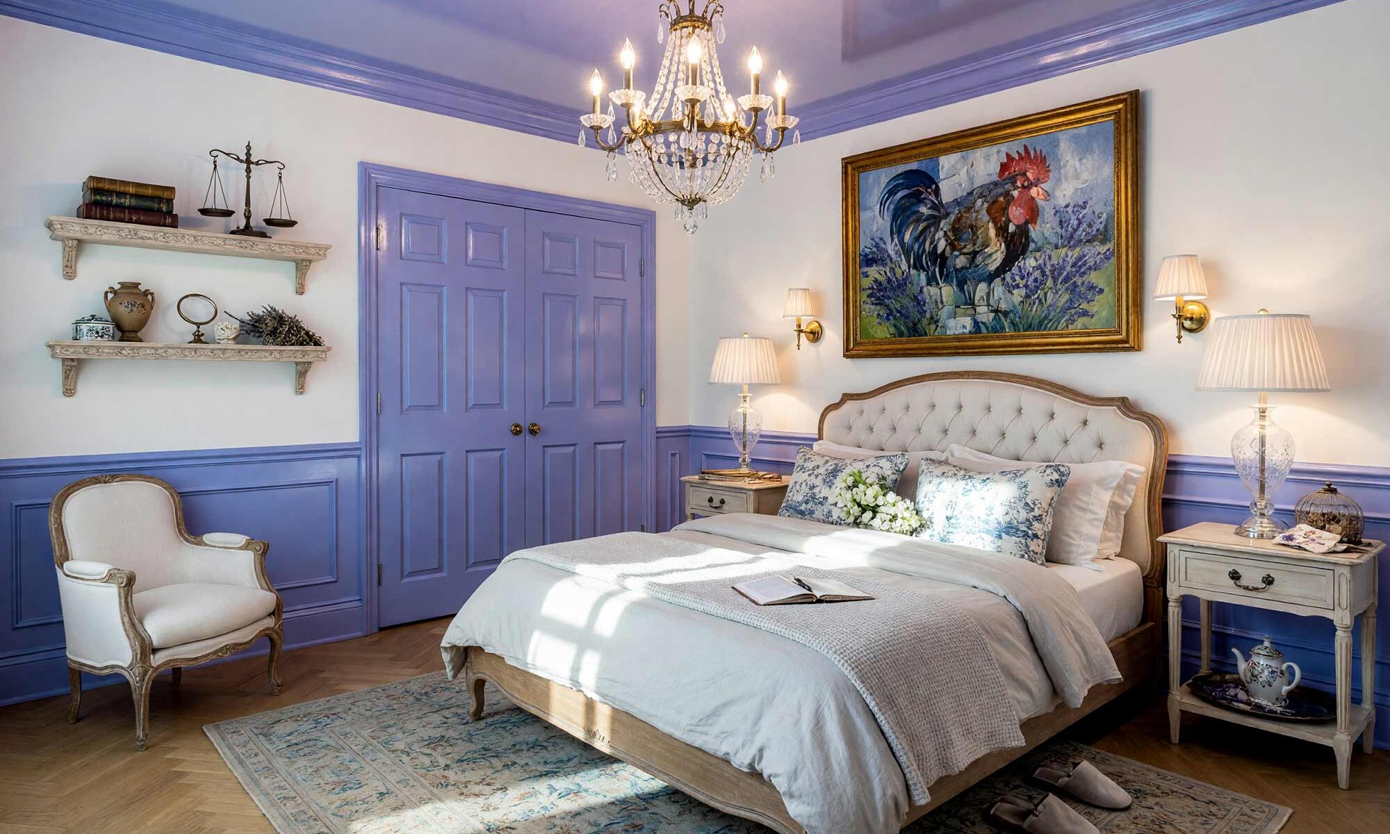 French Country Bedroom with White Walls and Periwinkle Ceiling and Wainscoting-1-FEATURE, french country primary bedroom cloud white walls periwinkle ceiling wainscoting high gloss interior paint white oak flooring. An exquisite French Country sanctuary in an upscale Scottsdale neighborhood pairing serene cloud white walls with a lustrous periwinkle lacquered ceiling and wainscoting. Featuring an authentic tufted linen bed dressed in rumpled luxury linens, vintage toile pillows, crystal chandelier illumination, and a curated French cubist painting. Dramatic sunlight enters through tall French doors, highlighting the artisanal herringbone floors and glistening painted woodwork to create a transcendent European escape.