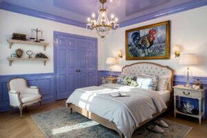 French Country Bedroom with White Walls and Periwinkle Ceiling and Wainscoting-1-FEATURE, french country primary bedroom cloud white walls periwinkle ceiling wainscoting high gloss interior paint white oak flooring. An exquisite French Country sanctuary in an upscale Scottsdale neighborhood pairing serene cloud white walls with a lustrous periwinkle lacquered ceiling and wainscoting. Featuring an authentic tufted linen bed dressed in rumpled luxury linens, vintage toile pillows, crystal chandelier illumination, and a curated French cubist painting. Dramatic sunlight enters through tall French doors, highlighting the artisanal herringbone floors and glistening painted woodwork to create a transcendent European escape.