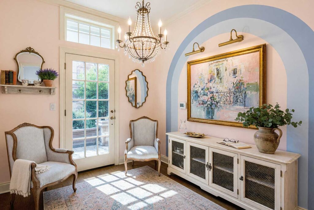 French Country Living Room Entry with Blush Pink Walls and Periwinkle Nested Arches-2, french country living room blush pink walls periwinkle painted wall graphics soft white ceiling french oak flooring. An elegant entryway in a Scottsdale neighborhood featuring blush pink walls, a soft white ceiling, and three nested periwinkle concentric arches. Complete with a distressed credenza, scalloped mirror, fresh eucalyptus pitcher, and wire-brushed French oak floors. High-contrast natural sunlight produces bold shadows throughout, capturing the timeless romance and curated beauty of French provincial design. graphic wall, wall graphic design, painted wall art, accent wall graphics