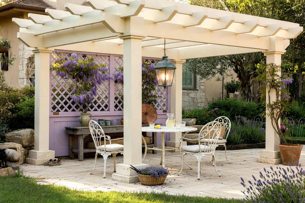 French Country Pergola with Warm Cream Posts and Lavender Trellis-1, french country pergola warm cream posts wisteria lavender trellis pergola paint colors. Situated in a custom Scottsdale courtyard, this rustic retreat pairs semi-gloss warm cream timber posts with a wisteria lavender trellis panel and matching roof slats. Distressed iron bistro seating, tumbled limestone, and fresh-cut lavender thrive beneath dynamic golden-hour light and deep shadows. pergola paint colors, painted pergola ideas, painting an outdoor pergola, painting backyard ramada, pergola color ideas, pergola color schemes, what color to paint a pergola