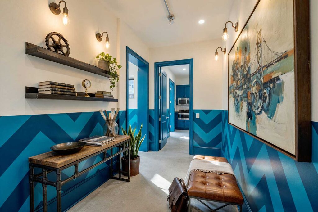 Industrial Loft Hallway with Cream Walls and Two-Tone Teal Blue Chevron Wainscot-2, industrial loft hallway cream walls teal blue painted wall graphics cream ceiling polished concrete flooring. A striking urban corridor in a Scottsdale neighborhood featuring cream walls, a cream ceiling, and a repeating diagonal chevron wainscot graphic in vivid teal blue. Outfitted with a reclaimed wood console, exposed-bulb sconces, a distressed leather bench, and polished concrete floors. Bold, directional daylight casts deep shadows, emphasizing the graphic precision of this modern loft transition space. graphic wall, wall graphic design, painted wall art, accent wall graphics