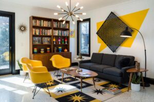 Living Room with Canary Yellow Speech-Balloon Burst Halftone and Perforated Aluminum Accent Wall-1, mid-century kitsch living room paper white walls primary canary accent wall halftone perforated aluminum signal black baseboards polished terrazzo floors painting creative wall treatments. A playful retro retreat in a Scottsdale neighborhood featuring a charcoal sectional sofa, a sputnik chandelier, packed mid-century bookshelves with action figures, and a dynamic atomic starburst area rug. The space is energized by an oversized yellow speech-balloon burst and a perforated aluminum accent panel casting intricate shadow patterns. Strong sunlight streams through large glass doors, highlighting the glossy signal black trim and the vibrant comic-book aesthetic. painting creative wall treatments, creative wall treatments, mixed material wall treatments, dimensional painted walls, textured accent wall ideas, metal accent wall ideas