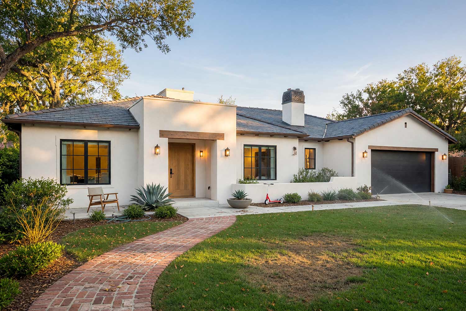 Modern Spanish Revival with Soft White Stucco Black Trim and Natural Oak Door-2, spanish revival exterior paint colors soft chalk white stucco pitch black window frames natural white oak door brick walkway front facade. A custom estate in a Scottsdale neighborhood featuring freshly painted stucco, glossy hand-finished trim, and elevated exterior painting curb appeal. A natural white oak door and pitch black window frames pair with soft chalk white stucco beneath a slate charcoal roof. A curving brick path, lush green lawn, mid-century entry seating, and desert sunset lighting complete this Palm Springs-inspired California Spanish exterior. santa barbara style paint colors, spanish hacienda exterior paint colors, mediterranean revival exterior colors, stucco house paint colors, terracotta roof house colors