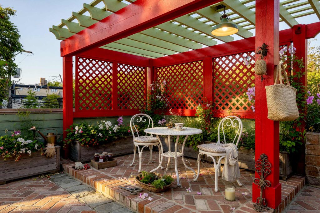 Pergola with Poppy Red Beams and Pistachio Green Overhead Slats-4-FEATURE, cottagecore backyard pergola poppy red posts pistachio green slats trellis pergola paint colors. Set within an upscale Scottsdale neighborhood, this vibrant shade structure showcases fresh semi-gloss poppy red posts paired with airy pistachio green slats and a matching trellis. An antique herringbone brick floor hosts a vintage café set beneath crisp dappled shadows, complemented by cut herbs and potted strawberries during golden hour. pergola paint colors, painted pergola ideas, painting an outdoor pergola, painting backyard ramada, pergola color ideas, pergola color schemes, what color to paint a pergola