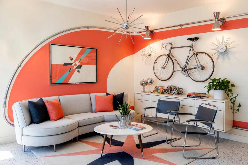 Retro Atomic Living Room with Cream Walls and Coral Curve with Polished Chrome Tubing-12, retro atomic living room vanilla cream walls atomic coral banking curve polished chrome tubing marshmallow baseboards terrazzo floors painting creative wall treatments. A kinetic mid-century lounge in a Scottsdale neighborhood featuring a curved sectional sofa, chrome sling chairs, terrazzo flooring, and an atomic coral banking curve accented with bent chrome tubing. Afternoon sun beams through patio doors, throwing sharp shadows that highlight the shiny marshmallow trim, starburst wall clock, and retro velodrome styling. painting creative wall treatments, creative wall treatments, mixed material wall treatments, dimensional painted walls, textured accent wall ideas, metal accent wall ideas