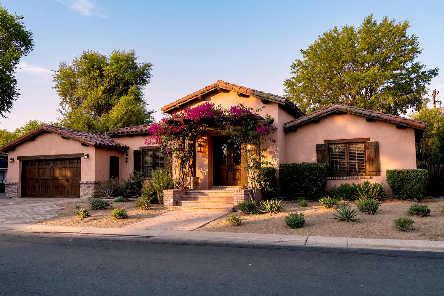 Santa Barbara Home with Dusty Desert Rose Walls and Dark Chocolate Trim-1, santa barbara exterior paint colors with dusty desert rose stucco dark chocolate trim red terracotta roof and paved concrete front facade. A custom Paradise Valley home pairing mineral matte plaster with semi-gloss millwork and classic red terracotta tile. Terraced planters, a brass watering can, an angled push-broom, and glowing amber coach lights complete the warm sunset curb appeal. santa barbara style paint colors, spanish hacienda exterior paint colors, mediterranean revival exterior colors, stucco house paint colors, terracotta roof house colors