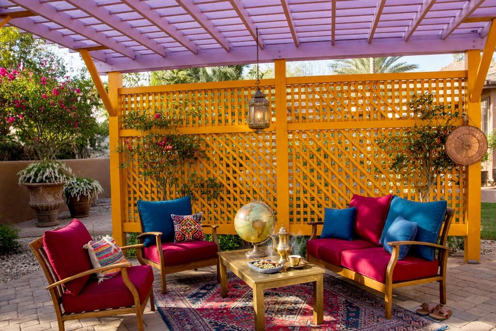 Silk Road Pergola with Saffron Framing and Orchid Slats-1, wanderlust silk road pergola saffron timber orchid slats trellis pergola paint colors. Located at an upscale Scottsdale residence, this Silk Road terrace pairs semi-gloss saffron posts and diamond lattice with dreamy orchid roof slats. Sandstone flagstones, a hand-woven rug, vintage globe, and glowing brass lantern shine under intense golden-hour shadows. painting a cedar pergola, ramada paint colors, trellis paint colors, backyard shade structure colors, best paint for an outdoor pergola