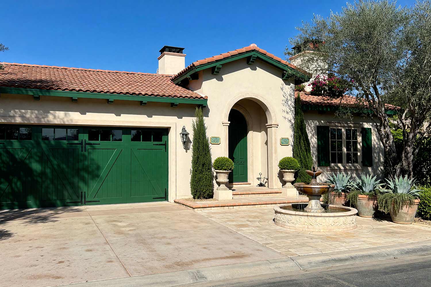 Spanish Revival Arcadia House with Cream Stucco and Bottle Green Shutters-3, spanish revival exterior paint colors buff cream stucco bottle green shutters blackout black rails concrete driveway front facade. A custom estate in a Scottsdale neighborhood featuring freshly painted stucco, glossy hand-finished trim, and elevated exterior painting curb appeal. Shiny bottle green shutters and an arched door accent buff cream stucco beneath burnt-orange terracotta roof tiles. An asymmetrical Cantera stone fountain, formal clipped boxwoods, and mature olive trees complete the traditional Spanish design under dynamic golden hour illumination. santa barbara style paint colors, spanish hacienda exterior paint colors, mediterranean revival exterior colors, stucco house paint colors, terracotta roof house colors