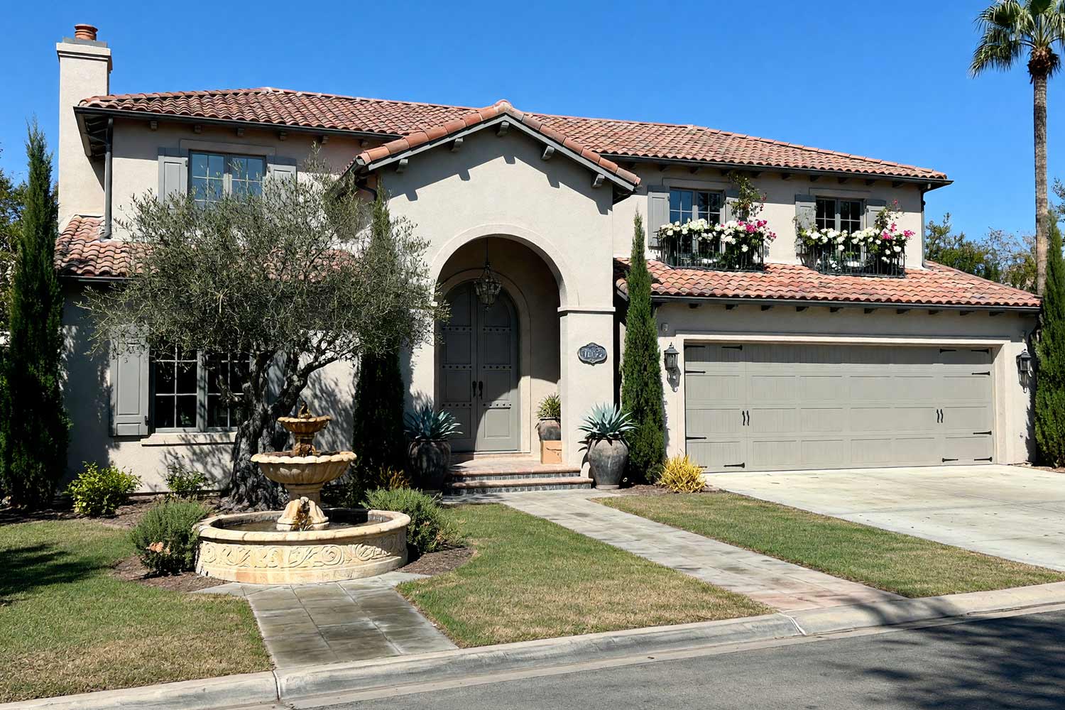 Spanish Revival Home with Pale Greige Stucco and Gray Shutters-3, spanish revival exterior paint colors pale french greige stucco storm-cloud gray shutters blackened slate rails concrete driveway front facade. A custom estate in a Scottsdale neighborhood featuring freshly painted stucco, glossy hand-finished trim, and elevated exterior painting curb appeal. Shiny storm-cloud gray shutters and an arched door pair with pale French greige stucco beneath russet terracotta barrel tiles. An asymmetrical Cantera stone fountain, crystal-drop loggia chandelier, and mature olive trees complete the Hollywood glam aesthetic under dramatic twilight light. santa barbara style paint colors, spanish hacienda exterior paint colors, mediterranean revival exterior colors, stucco house paint colors, terracotta roof house colors