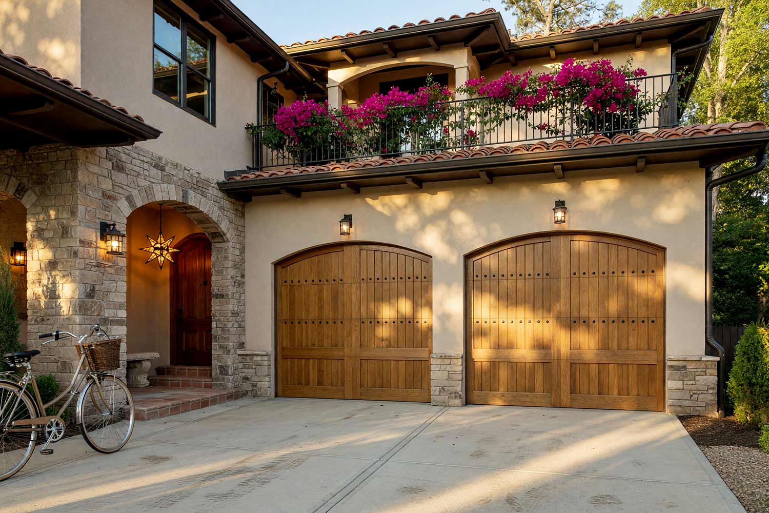 Spanish Revival Home with Warm Stucco Charcoal Trim and Natural Oak Garage Doors-2, spanish revival exterior paint colors warm limestone stucco deep charcoal trim natural white oak garage doors concrete driveway front facade. A custom estate in a Scottsdale neighborhood featuring freshly painted stucco, glossy hand-finished trim, and elevated exterior painting curb appeal. Two arched natural white oak garage doors pair with deep charcoal corbels and warm limestone stucco beneath weathered terracotta roof tiles. Stone entry arches, second-story loggia balconies, and minimalist iron lanterns create a peaceful Japandi-Spanish design under dynamic late-afternoon sunlight. santa barbara style paint colors, spanish hacienda exterior paint colors, mediterranean revival exterior colors, stucco house paint colors, terracotta roof house colors