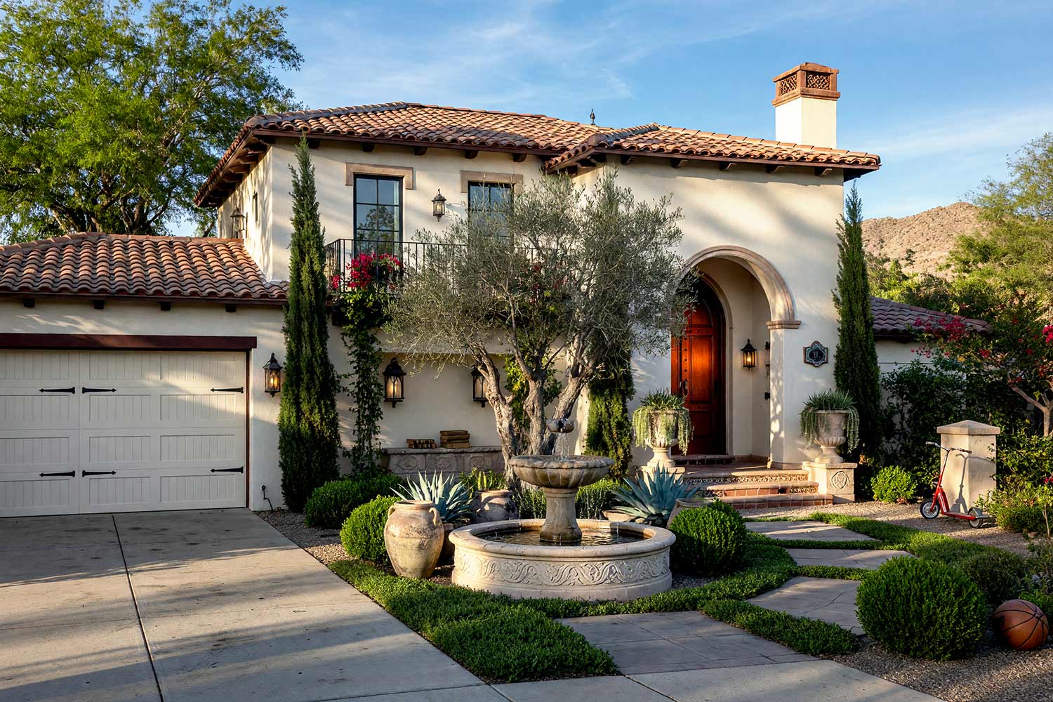 Spanish Revival House with Ivory Cream Stucco and Cordovan Oxblood Door-1, spanish revival exterior paint colors ivory cream stucco cordovan oxblood door raven black rails limestone walkway front facade. A custom estate in a Scottsdale neighborhood featuring freshly painted stucco, glossy hand-finished trim, and elevated exterior painting curb appeal. A glossy cordovan oxblood front door and matching canecillos corbels contrast against ivory cream stucco and deep mission terracotta roof tiles. An asymmetrical Cantera stone fountain, gothic-arch iron lanterns, and mature olive trees complete the scholarly dark academia aesthetic under dynamic golden hour illumination. spanish revival exterior paint colors, spanish style house paint colors, santa barbara exterior colors, modern mediterranean house paint, spanish colonial revival exterior colors