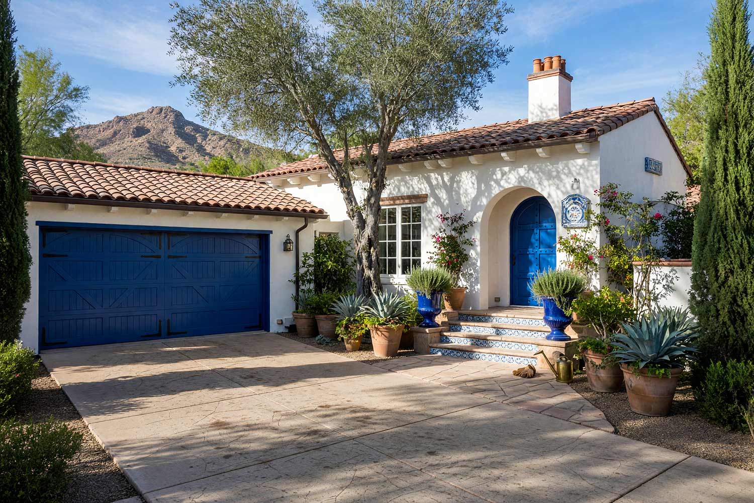 Spanish Revival Phoenix Home with White Stucco and Majolica Cobalt Doors-2, spanish revival exterior paint colors soft lime white stucco majolica cobalt door ink black lanterns concrete driveway front facade. A custom estate in a Scottsdale neighborhood featuring freshly painted stucco, glossy hand-finished trim, and elevated exterior painting curb appeal. French blue and cream Talavera tile stair risers lead to a saturated majolica cobalt front door beneath natural terracotta barrel tiles. An asymmetrical Cantera stone fountain bubbles softly alongside olive trees and vibrant pottery under dynamic golden hour illumination. spanish revival exterior paint colors, spanish style house paint colors, santa barbara exterior colors, modern mediterranean house paint, spanish colonial revival exterior colors