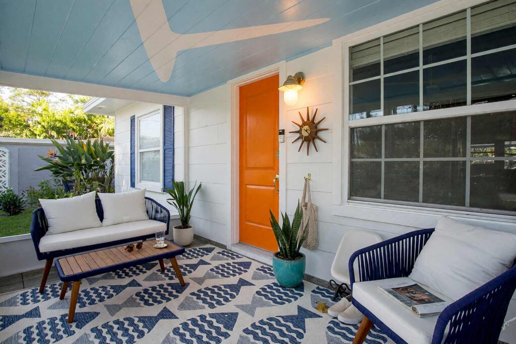 Mid-Century Entry Ceiling in Powder Blue with Pale Boomerangs and Tangerine Door-8, mid-century modern porch ceiling ideas powder blue boomerang shapes bright white fascia tangerine door. At a custom Scottsdale ranch, this covered entry pairs a semi-gloss powder blue ceiling and pale boomerangs with matte light gray siding. A brass starburst clock, molded fiberglass seating, and a tangerine door deliver playful retro curb appeal. Dynamic golden hour lighting sculpts deep shadows and brilliant highlights across the woodwork. porch ceiling paint ideas, patio ceiling color ideas, painted patio ceiling, striped porch ceiling, black porch ceiling, exterior ceiling paint colors, Southern porch ceiling colors, desert porch ceiling colors