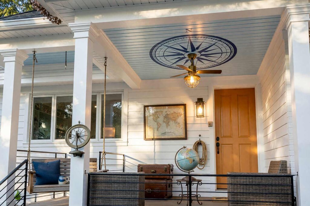 Porch Ceiling with Pale Haint Blue Ceiling with Painted Compass Medallion-2, pale haint blue porch ceiling ideas deep indigo compass medallion bright white columns persimmon door. This custom Scottsdale residence balances matte warm white siding with shiny semi-gloss painted woodwork. An overhead deep indigo compass-rose medallion pairs with brass nautical accents, a vintage globe, and antique travel trunks. Moody late-afternoon golden hour lighting sculpts deep shadows and radiant highlights across the haint blue ceiling. porch ceiling ideas, painted porch ceiling ideas, haint blue porch ceiling, front porch ceiling paint colors, covered patio ceiling paint, blue porch ceiling, haint blue paint colors
