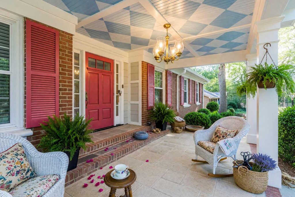 Porch Ceiling with Powder Blue Harlequin Pattern Bright White Trim and Raspberry Accents-8, grandmillennial porch ceiling ideas powder blue harlequin diamond pattern bright white trim raspberry door. This custom home in an upscale Scottsdale neighborhood balances matte light greige siding with shiny semi-gloss painted ceiling woodwork. The hand-painted powder blue harlequin diamond grid pairs with a glowing brass chandelier, vintage china, and lush boxwood accents. Dynamic late-afternoon golden hour lighting casts deep, dramatic shadows across the overhead structural beams. porch ceiling ideas, painted porch ceiling ideas, haint blue porch ceiling, front porch ceiling paint colors, covered patio ceiling paint, blue porch ceiling, haint blue paint colors
