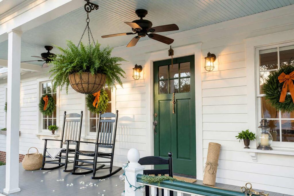 Veranda Ceiling with Pale Haint Blue Ceiling and White Walls and Hunter Green Door-3, pale haint blue porch ceiling ideas oyster white siding bright white columns hunter green door. Situated at a custom estate in a Scottsdale neighborhood, this Colonial veranda showcases a shiny semi-gloss pale haint blue ceiling against matte oyster white siding. A dark ceiling fan, lush hanging Boston fern, and painted rocking chair complete the traditional aesthetic. Moody late-afternoon golden hour sunlight creates sharp shadows and warm highlights overhead. porch ceiling ideas, painted porch ceiling ideas, haint blue porch ceiling, front porch ceiling paint colors, covered patio ceiling paint, blue porch ceiling, haint blue paint colors